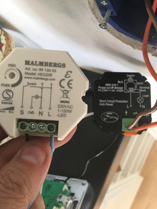 En hand håller en Malmbergs HD2200 dimmer bredvid en installerad DRS 315-1 dosdimmer mot en vit takyta.