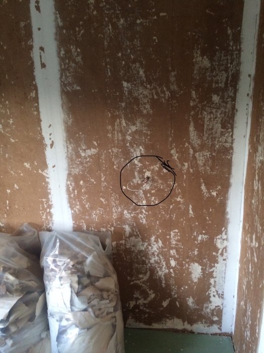 Skadad brun vägg med flagnande färg och en cirkel markerande en defekt, med säckar av rivningsmaterial framför.