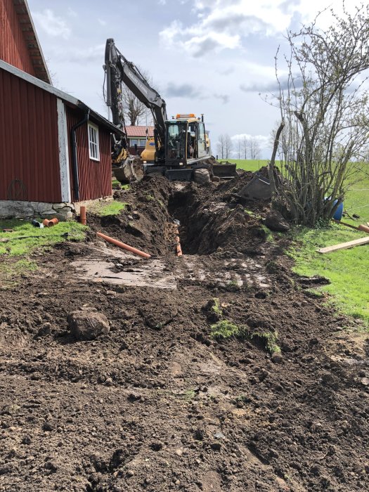 Grävmaskin arbetar i ett trångt och lerigt område bredvid en röd stuga.