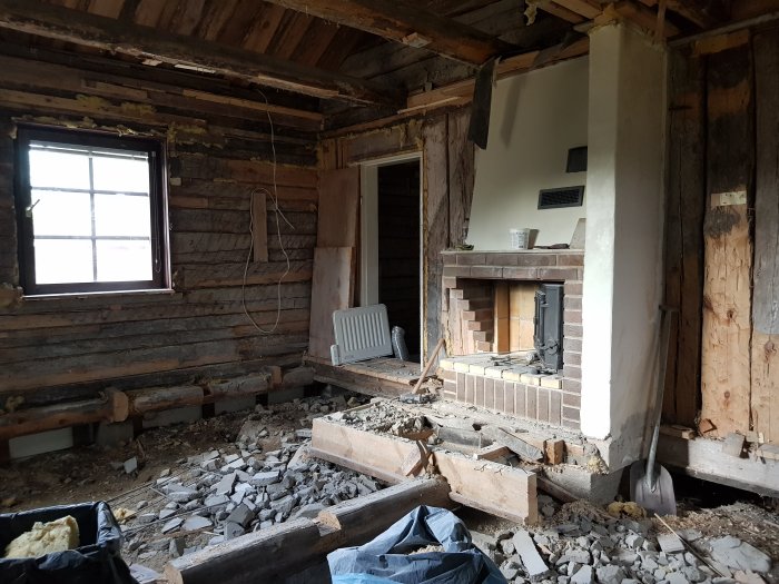 Inre renoveringsarbete på ett gammalt hus med exponerade trästockar, rivet golv och spis.
