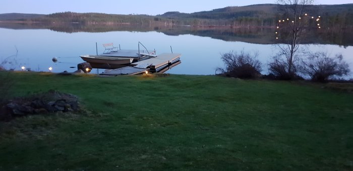 Alt: Skymning över en stilla sjö i Dalarna med en brygga och båt, omgiven av skog och en grön gräsmatta.