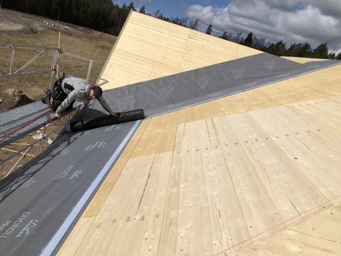 Person som lägger takduksmaterial på nybyggt trähus under konstruktion med synliga takstolar och byggställningar.