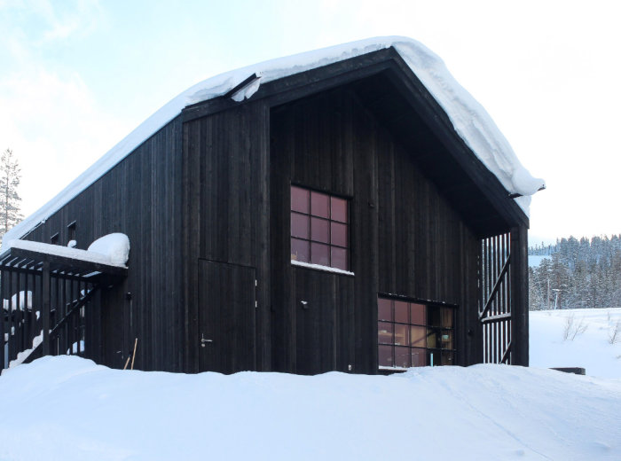 Svart trästuga med stora fönster mot en snötäckt mark och skog i bakgrunden.