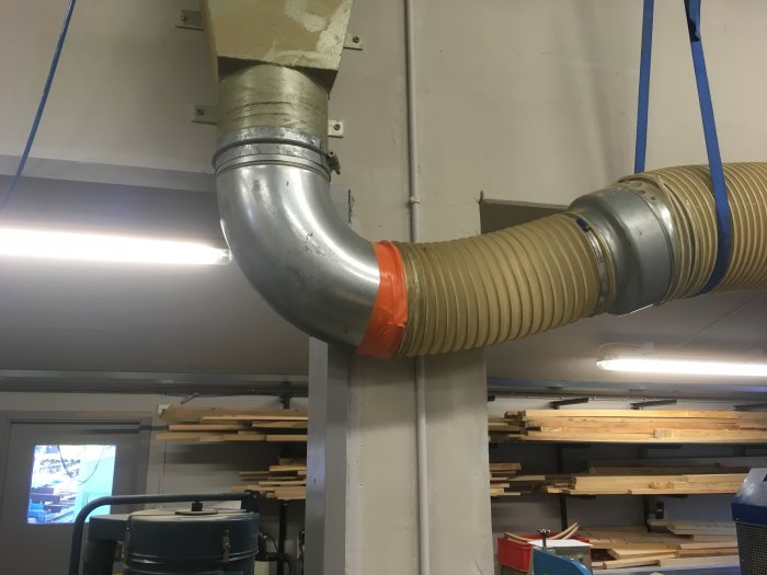 Flexibel avgasarm ansluten till fast ventilationssystem med orange tejp i ett snickeri.