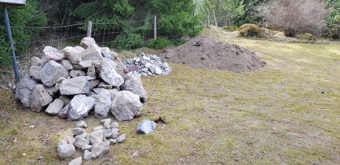 En hög med stora stenar och en grävd jordhög i en trädgård som visar arbete med stenspräckning och grävning.