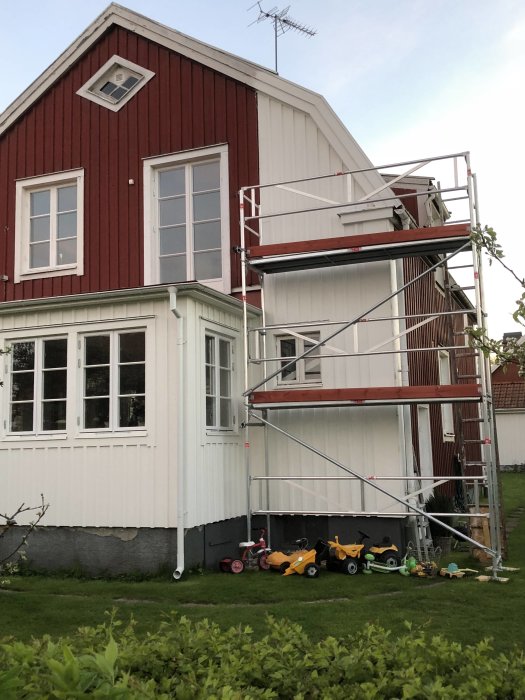 Rödvitt hus delvis målat med byggnadsställning framför, och leksaker spridda på gräset.