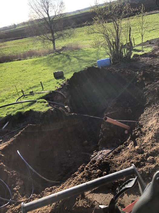 Grävd grop för byggprojekt med synliga elledningar och verktyg i en trädgård.