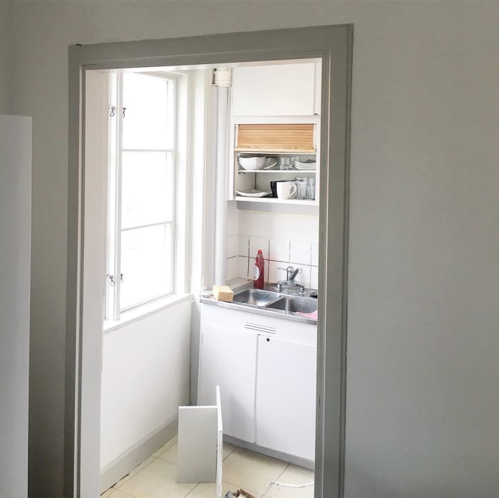 Vy genom en dörröppning in i ett litet kök med vita skåp, hyllor med porslin och ett fönster.
