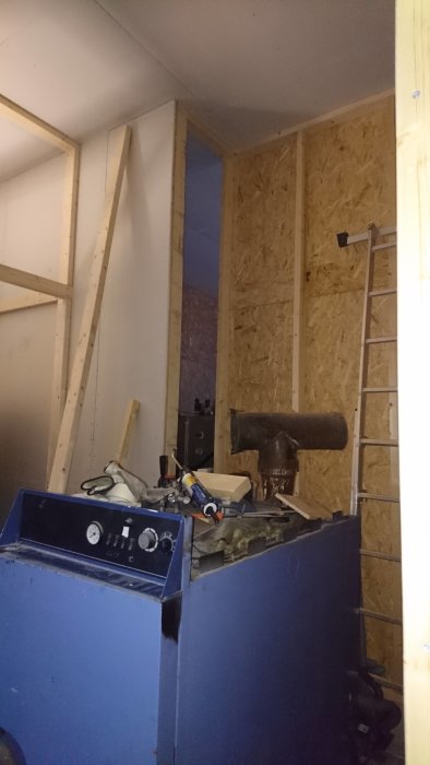 Byggarbetsplats med rör och verktyg framför OSB-väggar, stege till höger.