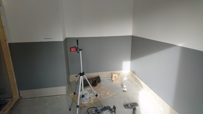 Omfärgat rum med halvfärdigt klinkergolv, byggmaterial och laseravståndsmätare på stativ.