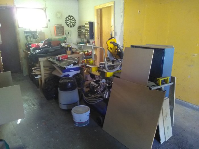 Rörigt garage med verktyg och material, slitet betonggolv och gula väggar i dagsljus.