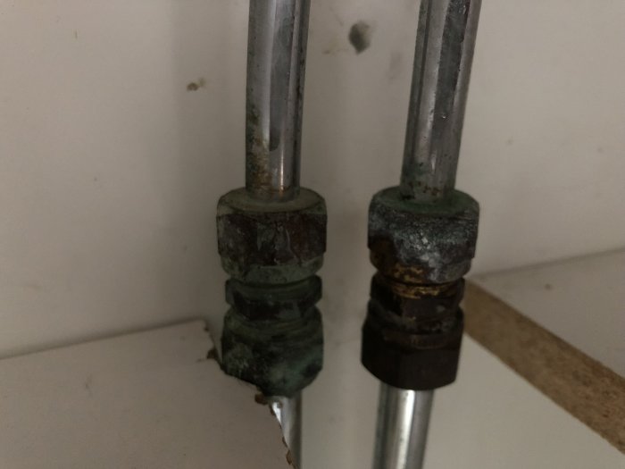 Äldre rostiga vattenrör och kopplingar mot en vit vägg, behov av byte inför kommodrenovering.