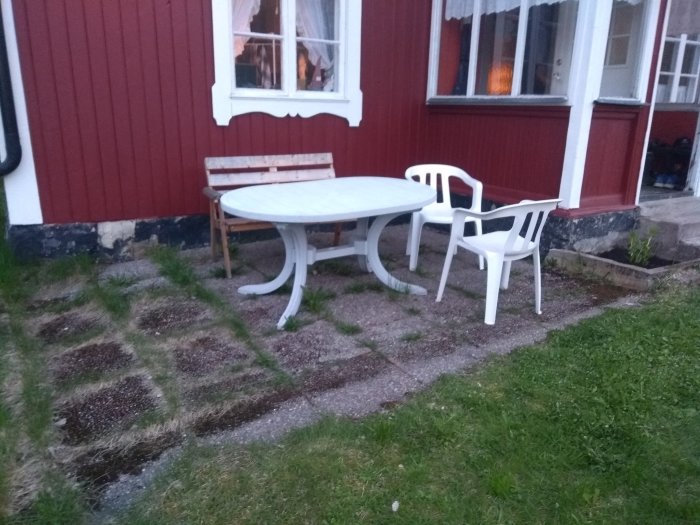 Gammal stenplattor med mossa vid husvägg under ett bord och stolar, gräs runtomkring.