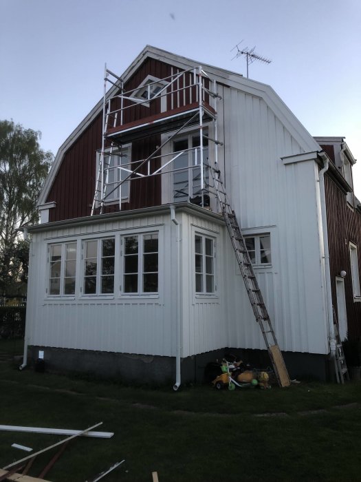 Byggnadsställning på verandatak av hus under renovering med ommålat virke och stege.