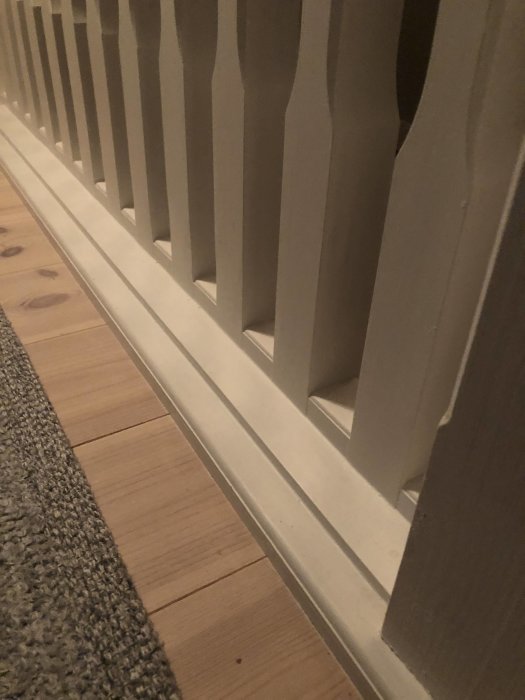 Detaljbild av ett nyligen byggt träräcke med vita spjälor och en bottenstycke, inomhus mot ett trägolv.