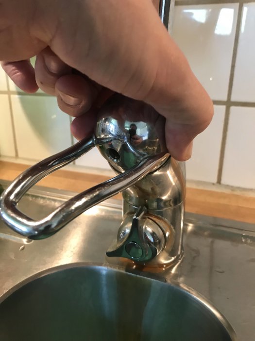 Hand försöker lyfta av handtaget på en köksblandare som sitter fast, med kakelvägg i bakgrunden.