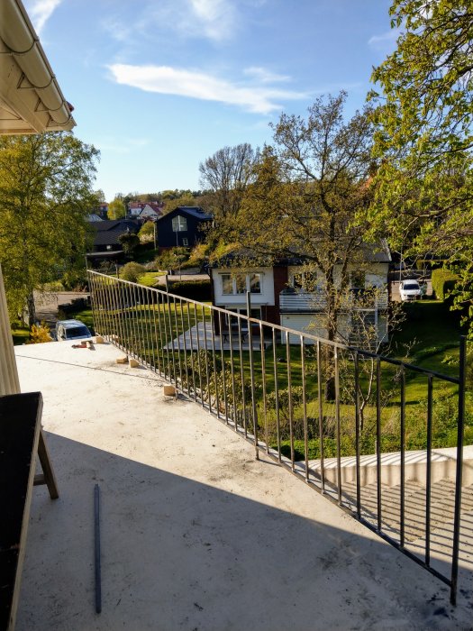 Vy från en ofärdig balkong med svarta räcken över ett bostadsområde med gröna träd och hus.