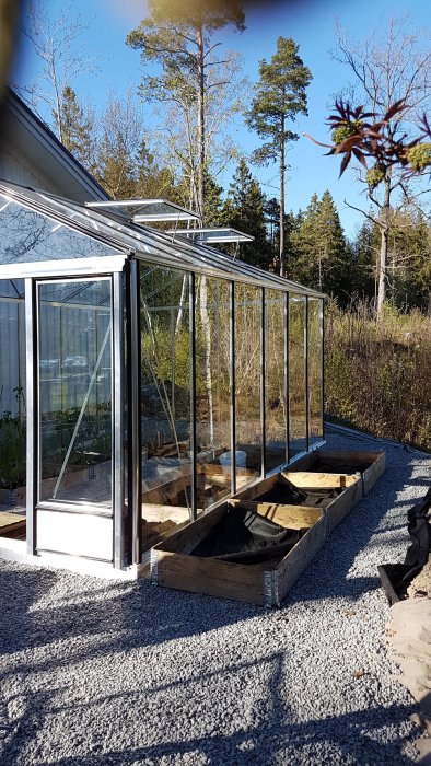 Nyligen uppfört växthus med öppna dubbeldörrar och pallkragar för plantering på grusbelagd yta, omgiven av natur.