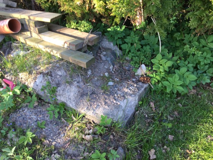 Tjock betongklump vid tomtkant, omgiven av gräsmatta och buskar, med träplankor ovanpå.