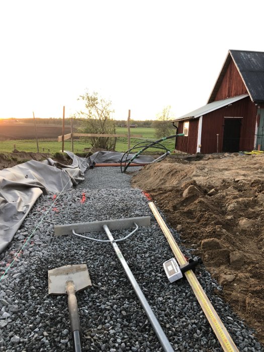 Byggarbetsplats med makadam, verktyg, nivåmätare och en grävd yta vid sidan under tidig kvällssol intill en röd lada.