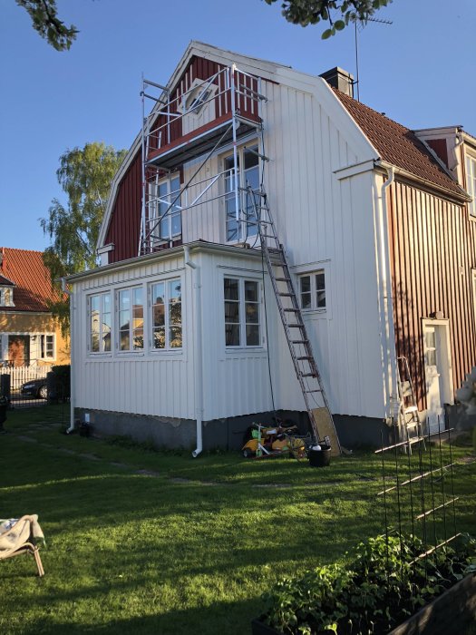 Ställning uppställd vid rödvit trävilla under renovering, med stege och byggmaterial i förgrunden.