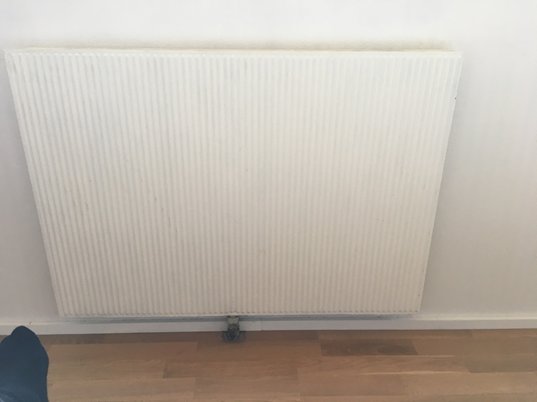 Vit radiator med synlig underliggande rörkoppling och en bit av ett golv.