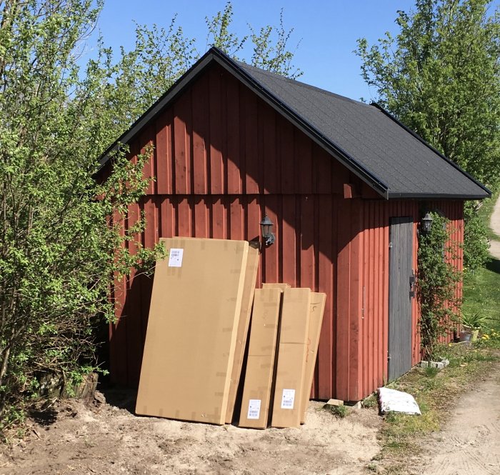 Levererade lådor med växthusdelar lutade mot en röd lada vid sidan av en grusväg.