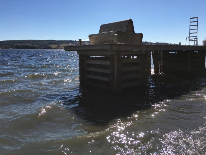 Brygga med intakt kista och skadat vågskydd vid vattnet, solljus på vågor.