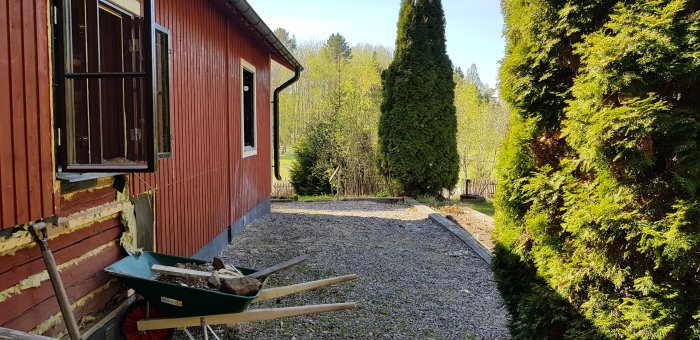 En röd husvägg med synlig isolering och öppet fönster, skottkärra med grus framför, omgiven av gröna träd och buskar.