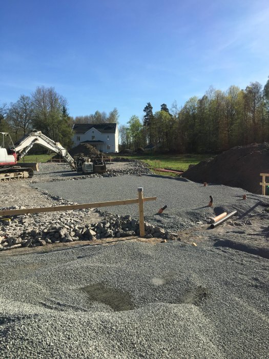 Grundarbete för byggnation med grus, grävmaskin och avmärkta punkter för stolpar mot hus i bakgrunden.