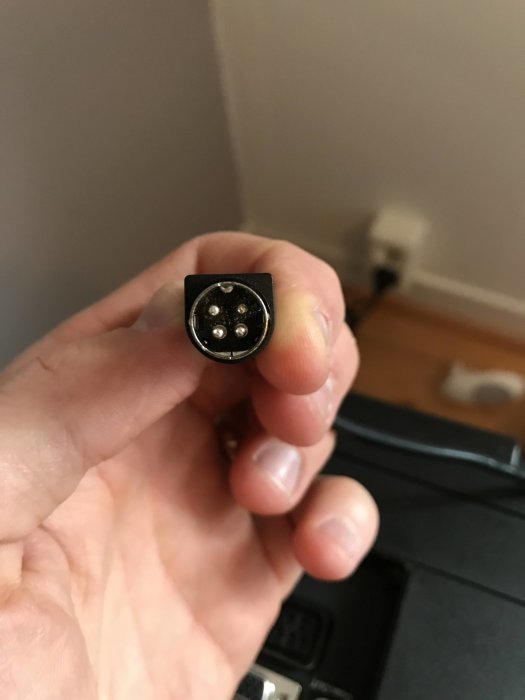 Hand håller en rund TV-kontakt med flera pinnar, mot en bakgrund av en vägg och golv.