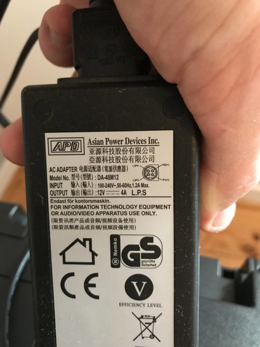 Hand håller en AC-adapter från Asian Power Devices med informationsetikett synlig.