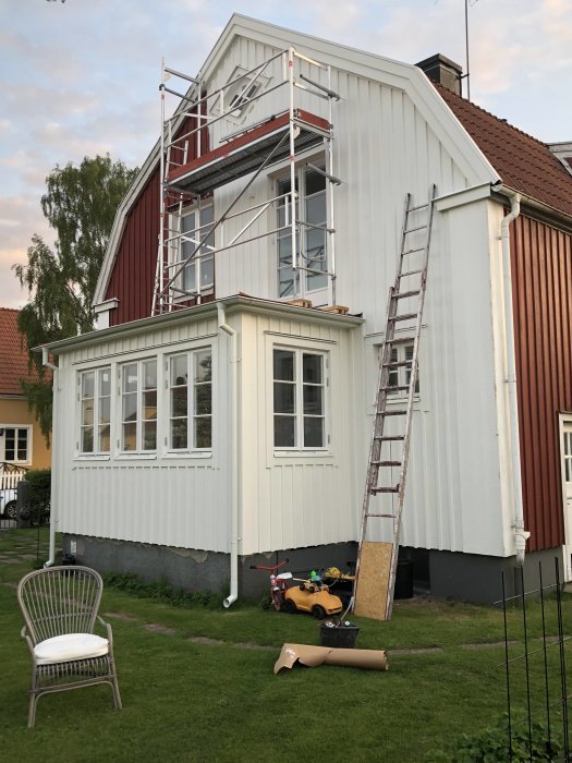 Ett hus med röda och vita sektioner och en ställning vid ena gaveln, verktyg och stege på marken.