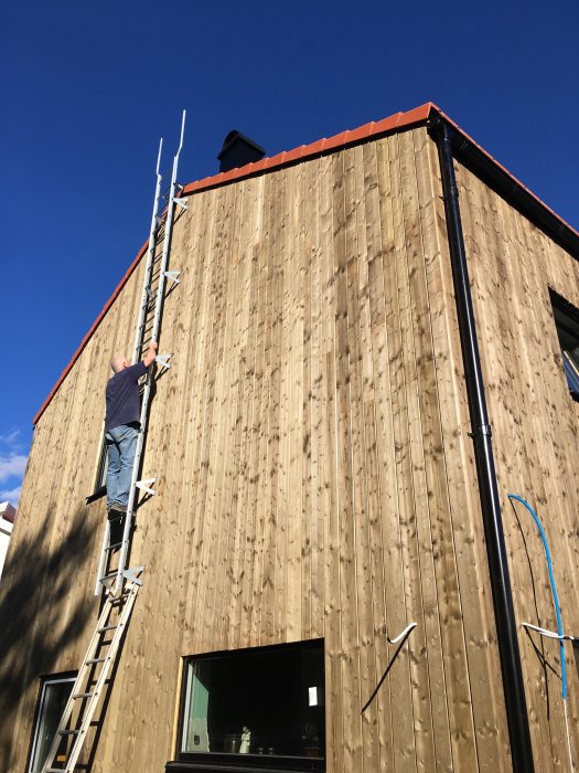 En person på en stege vid ett hus med träfasad behandlad med Herdins, under en klarblå himmel.