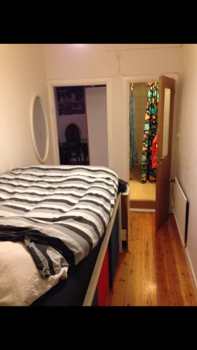 Före-bild av ett sovrum med säng, trägolv och öppen dörr till en annan del av huset.