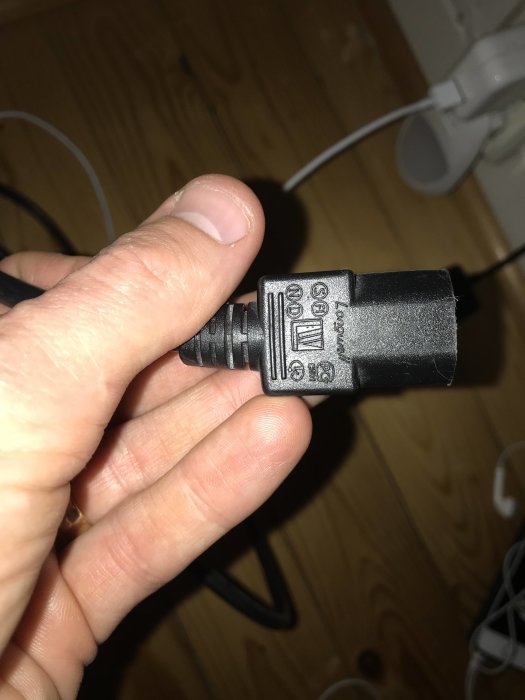 Hand håller en elektrisk transformatorplugg över ett trägolv med andra kablar i bakgrunden.