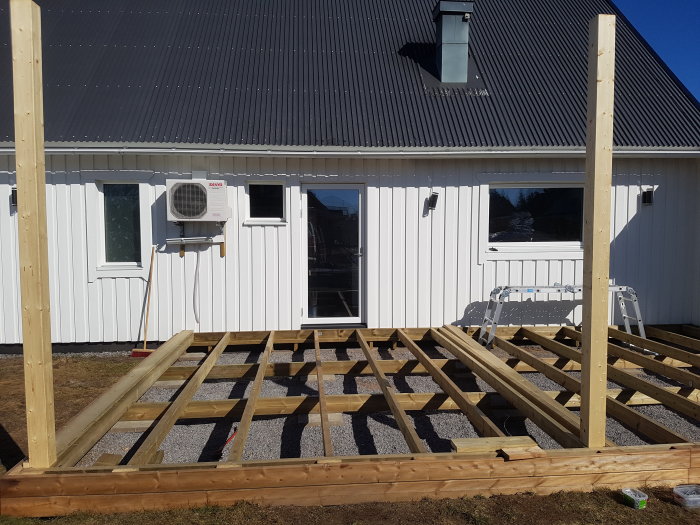 En oavslutad träkonstruktion för en uteplats intill en vit husfasad med dörr och fönster.
