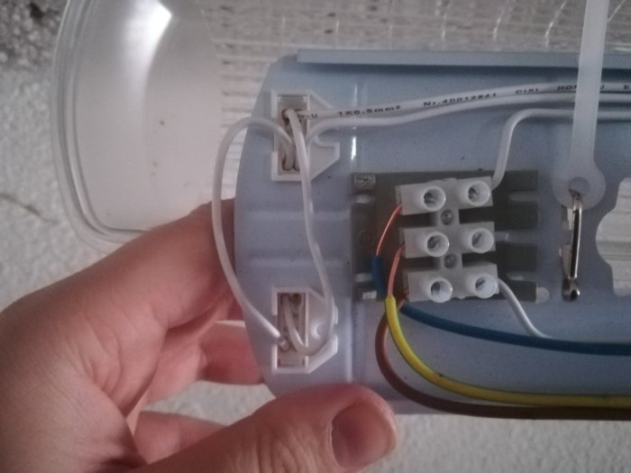 Hand som håller en öppen armatur med synligt illa skalade elektriska kablar och anslutningar inuti.