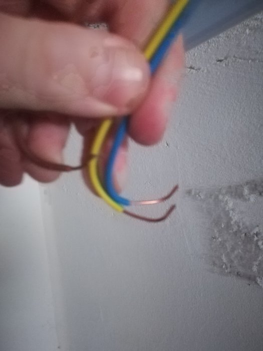 Hand håller felaktigt skalade elektriska kablar med synliga koppartrådar mot en vit vägg.