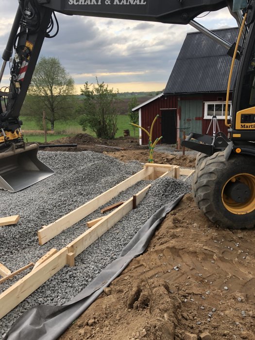 Förberedelse av grund med armeringsjärn och grus inför gjutning av en betongplatta nära en grävmaskin.