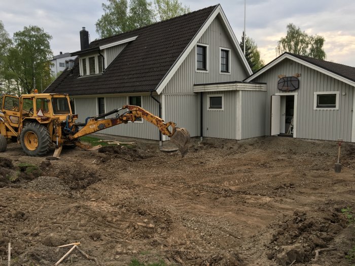 Gul grävmaskin vid arbete i trädgård framför vit husfasad med mörkt tak.