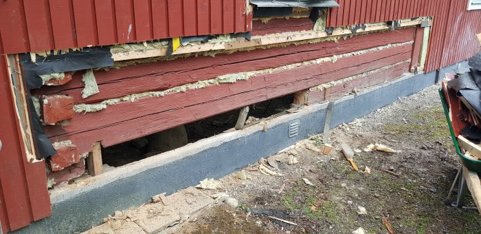 Del av träfasad på hus med synlig skada och isolering, understödd av provisoriskt stöd.