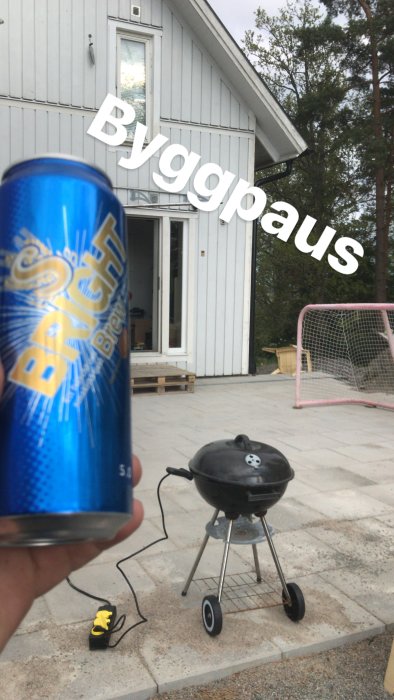 Hand håller en blå burk med etiketten 'Byggpaus' framför ett vitt hus med öppen dörr, en grill och fotbollsmål synliga.