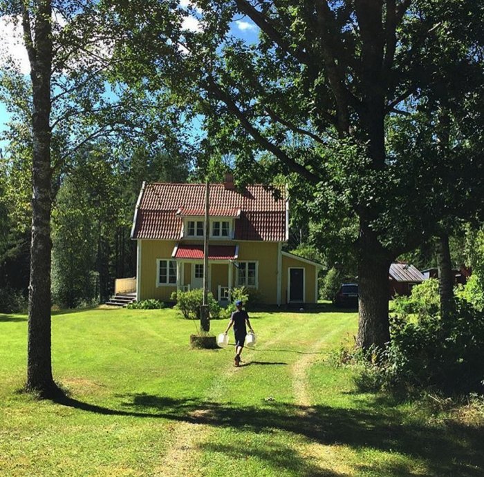 Person klipper gräset framför en gul villa på landet med röda fönsterluckor, omgiven av träd i soligt väder.