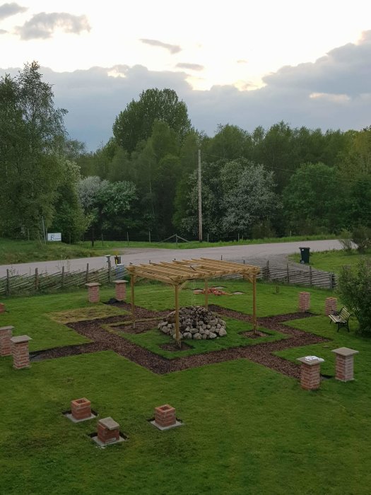 En ofärdig trädgårdsanläggning med träpelare, vildvin planteringar och en nybyggd pergola mot en grönskande bakgrund.