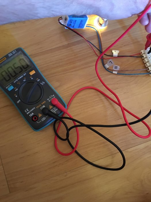 Digital multimeter ansluten till en kondensator för att mäta spänningen, med testerledningar på trägolv.