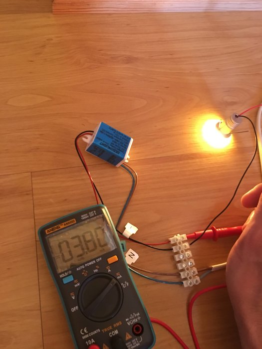Elektrisk mätning av spänning med multimeter och en tänd lampa, kopplingsklämmor och kablar på trägolv.