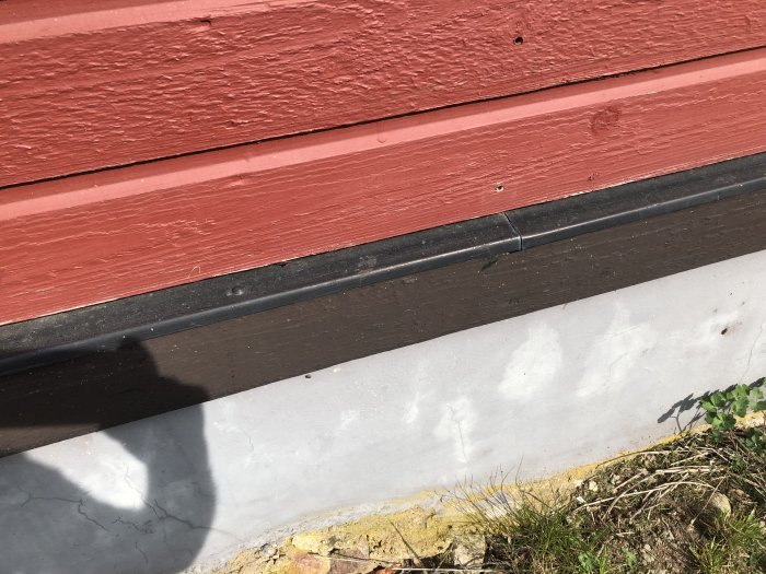 En metallplåt monterad mellan röd träpanel och brun träbräda ovanför betonggrund på ett hus.