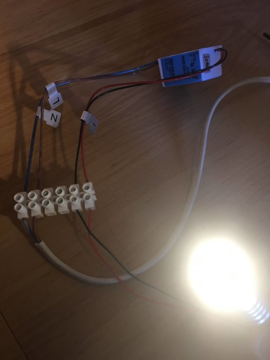 Elinstallation med öppna kablar, klämmor och en tänd glödlampa på ett trägolv.