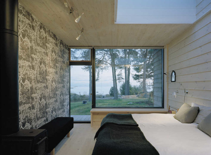 Modernt sovrum med stort fönster ned till golvet med utsikt över trädgård och hav.